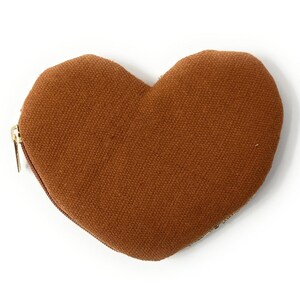 May include: A heart-shaped, brown fabric coin purse with a gold zipper. The purse is made of a textured material and is set against a white background. The design is simple and elegant.