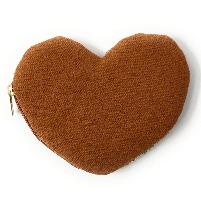 May include: A heart-shaped, brown fabric coin purse with a gold zipper. The purse is made of a textured material and is set against a white background. The design is simple and elegant.