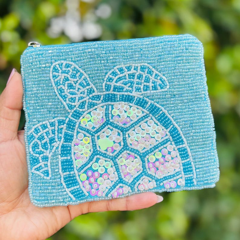 K&ouml;nnte beinhalten: Eine hellblaue, perlenbesetzte Tasche mit Rei&szlig;verschluss. Die Tasche hat ein wei&szlig;es und schillerndes Pailletten-Design einer Meeresschildkr&ouml;te. Der Panzer der Meeresschildkr&ouml;te ist mit schillernden Pailletten verziert. Die Tasche wird in einer Hand gehalten.
