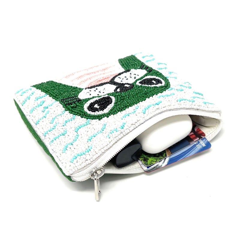 K&ouml;nnte beinhalten: Eine wei&szlig;e, perlenbesetzte Tasche mit einem gr&uuml;nen Hundegesicht-Design. Die Tasche ist offen und zeigt einen wei&szlig;en Gegenstand, einen schwarzen Gegenstand und eine Karte. Der Rei&szlig;verschluss ist silberfarben.