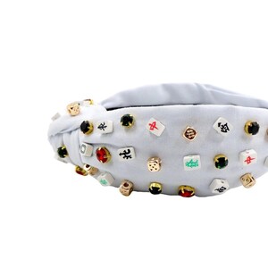 A white headband with a twisted design, adorned with various colorful gemstone studs and Mahjong tile charms, conveying a playful and eclectic style.