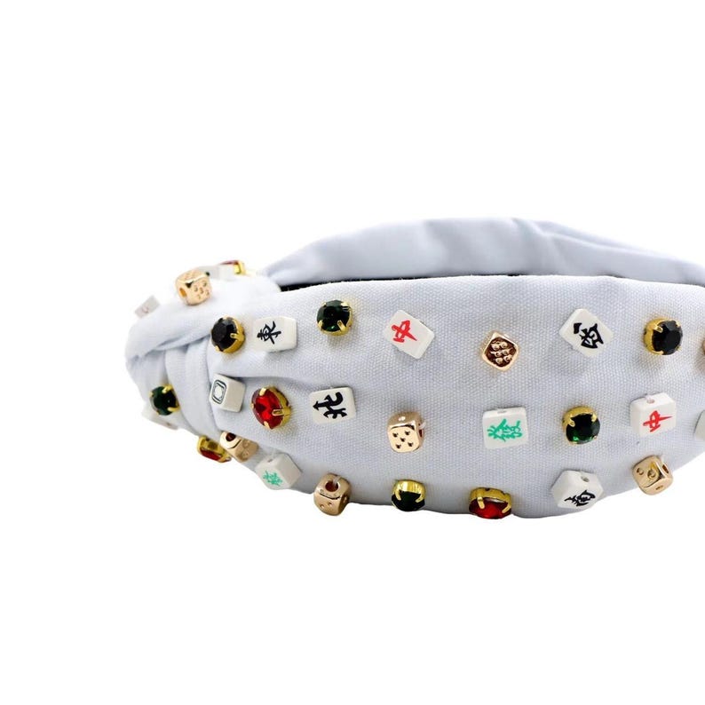 A white headband with a twisted design, adorned with various colorful gemstone studs and Mahjong tile charms, conveying a playful and eclectic style.
