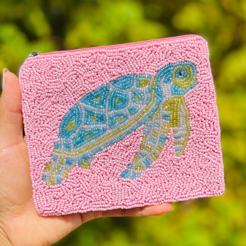 K&ouml;nnte beinhalten: Rosa Perlenbeutel mit einem Seeschildkr&ouml;ten-Design. Die Schildkr&ouml;te ist aus blauen, gelben und wei&szlig;en Perlen gefertigt. Der Beutel hat einen Rei&szlig;verschluss und wird in einer Hand gehalten.