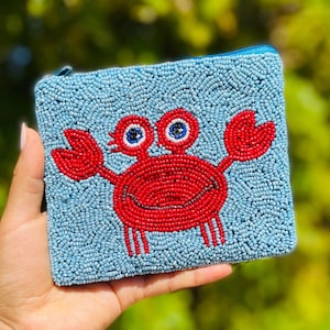 K&ouml;nnte beinhalten: Eine quadratische, perlenbesetzte Tasche mit hellblauem Hintergrund und einem roten Krebsdesign. Der Krebs hat ein l&auml;chelndes Gesicht und blau-wei&szlig;e Augen. Die Tasche hat einen blauen Rei&szlig;verschluss.