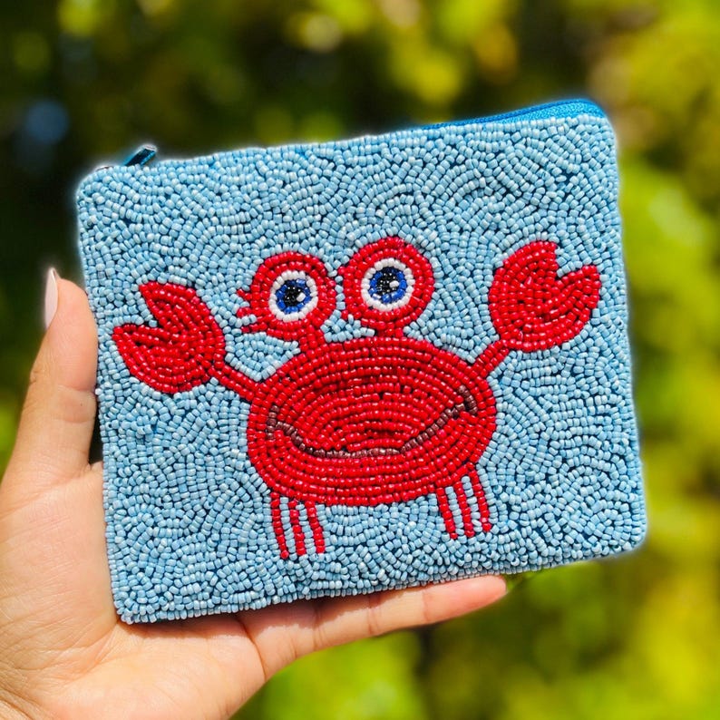 K&ouml;nnte beinhalten: Eine quadratische, perlenbesetzte Tasche mit hellblauem Hintergrund und einem roten Krebsdesign. Der Krebs hat ein l&auml;chelndes Gesicht und blau-wei&szlig;e Augen. Die Tasche hat einen blauen Rei&szlig;verschluss.