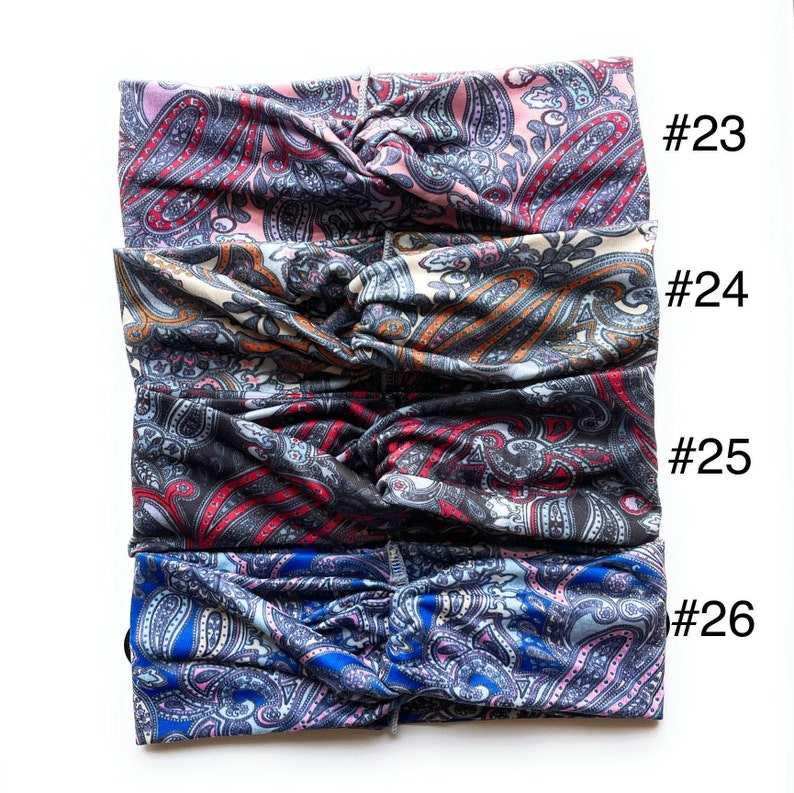 May include: Four different patterned headbands stacked on top of each other. The top headband is pink with a paisley pattern. The second headband is beige with a paisley pattern. The third headband is black with a paisley pattern. The bottom headband is blue with a paisley pattern. The headbands are numbered #23, #24, #25, and #26.