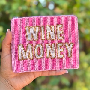 May include: A pink beaded clutch with the words "WINE MONEY" in gold and white beads. The clutch has a striped pattern in shades of pink. The background is blurred green foliage.