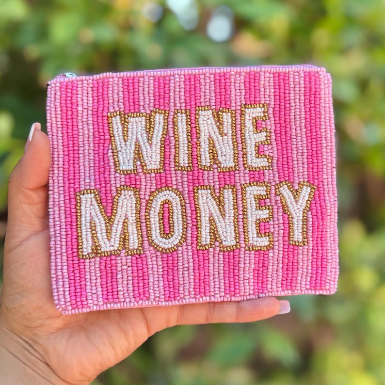 May include: A pink beaded clutch with the words "WINE MONEY" in gold and white beads. The clutch has a striped pattern in shades of pink. The background is blurred green foliage.