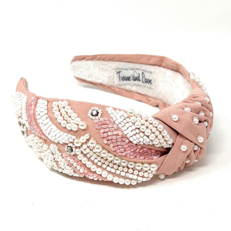 May include: A pink knotted headband with a white and pink beaded design. The headband is decorated with pearls and sequins. The headband has a white fabric lining and the text "Twine and Love" is printed on the lining.