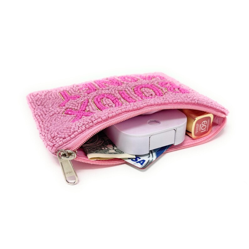 May include: Pink beaded pouch with the word "Juicy" embroidered in pink. The pouch is open, revealing a white case, lipstick, cash, and a credit card. The zipper is silver.