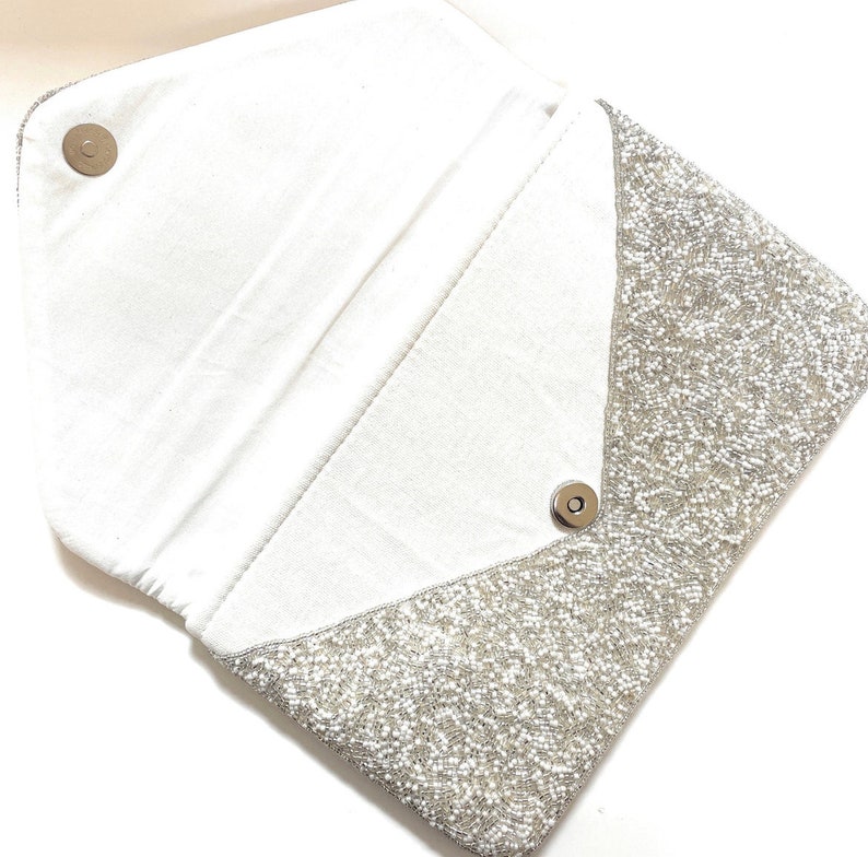 Op de afbeelding: Een zilveren clutch met parelversiering, een witte voering van stof en een drukknopsluiting.