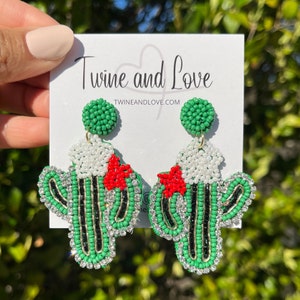 May include: Green and red beaded cactus earrings with a white beaded flower and rhinestones. The earrings are on a white card with the text "Twine and Love" and "twineandlove.com".