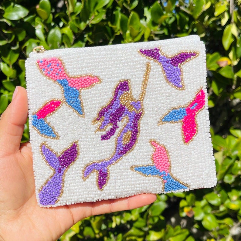 K&ouml;nnte beinhalten: Wei&szlig;e, perlenbesetzte Tasche mit goldenem Rei&szlig;verschluss, mit einer Meerjungfrau und mehreren Meerjungfrauenschwanz-Designs in Pink, Blau und Lila. Die Tasche wird in einer Hand vor einem Hintergrund aus gr&uuml;nem Laub gehalten.