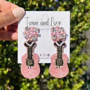 May include: Pink beaded guitar earrings with a rhinestone flower detail. The earrings are on a white card with the text "Twine and Love" and social media icons.
