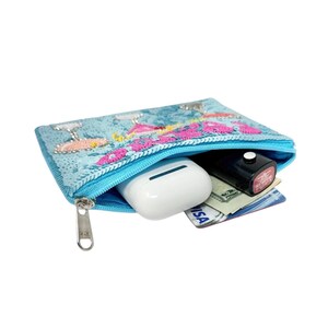 May include: A light blue zippered pouch with a floral design in pink and white sequins. The pouch is open, revealing a pair of white earbuds, a black lipstick, cash, and a credit card.