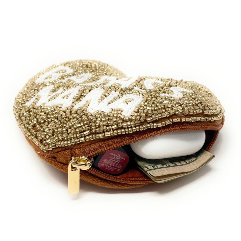 May include: A heart-shaped, beaded coin purse with the words "Bad Nana" in white. The purse is gold and brown with a gold zipper and contains a lipstick, a white earbud case, and folded cash.