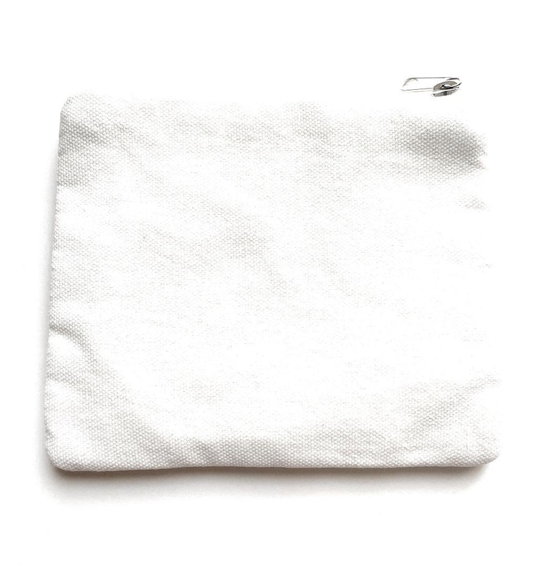 K&ouml;nnte beinhalten: Ein wei&szlig;es Canvas-Etui mit einer silbernen Sicherheitsnadel, die an der oberen rechten Ecke befestigt ist. Das Etui ist rechteckig und hat eine strukturierte Oberfl&auml;che. Der Hintergrund ist wei&szlig;.