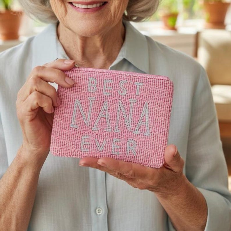 May include: A pink beaded clutch with the words "BEST NANA EVER" in light gray lettering. The clutch is rectangular and has a zipper closure. The background is a light blue shirt.