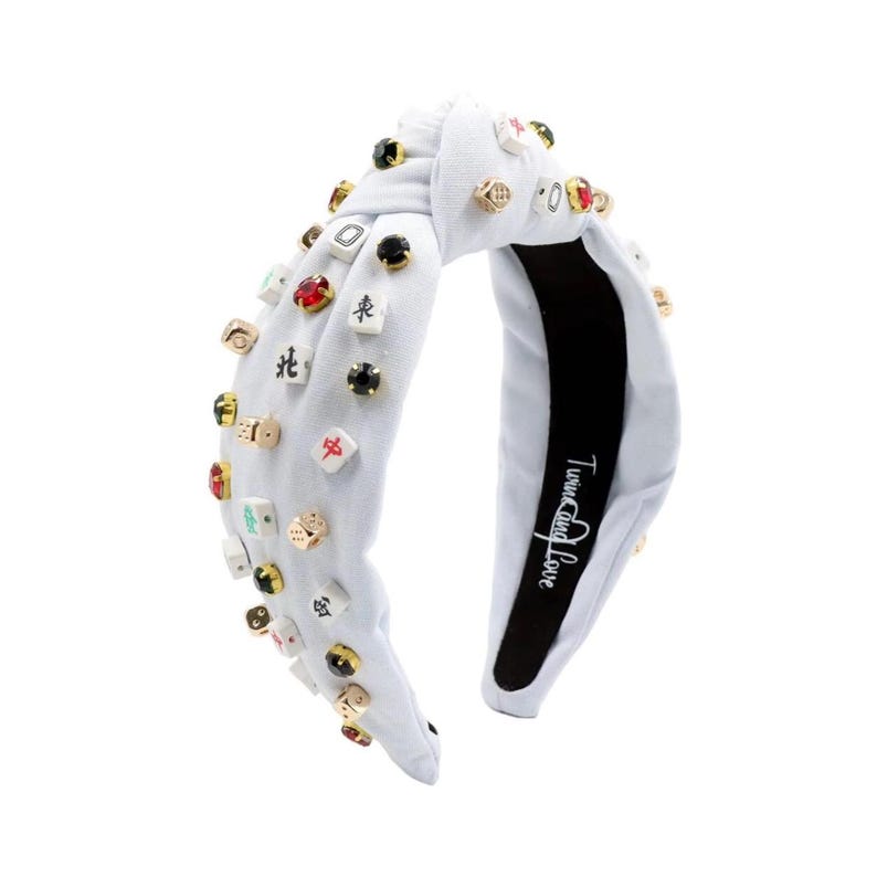 A white headband with a twisted design, adorned with various colorful gemstone studs and Mahjong tile charms, conveying a playful and eclectic style.