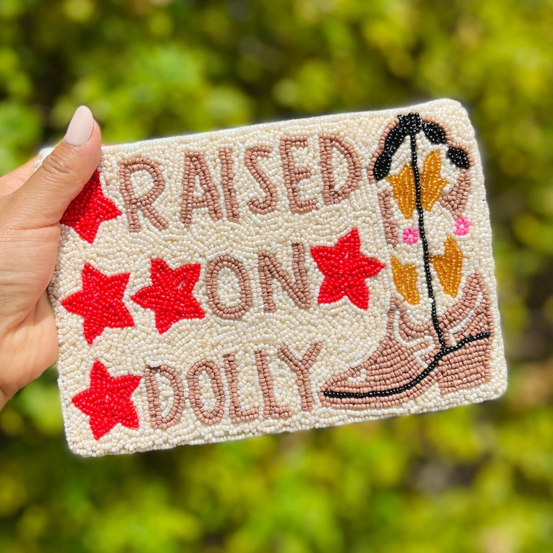 May include: A white beaded clutch bag with the text "RAISED ON DOLLY" and a brown cowboy boot design. The text is in brown beads and the stars are red.