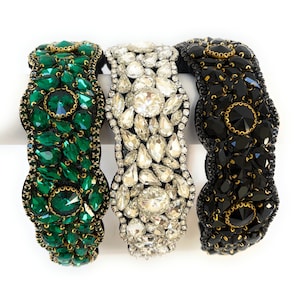 May include: Three headbands with different colored rhinestones. The headband on the left has green rhinestones, the middle headband has clear rhinestones, and the headband on the right has black rhinestones.