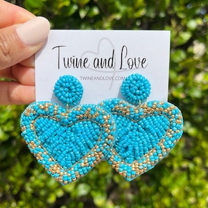 May include: Turquoise blue heart-shaped beaded earrings with gold accents. The earrings are large and dangle. The brand name "Twine and Love" and website "twineandlove.com" are printed on a white card behind the earrings.