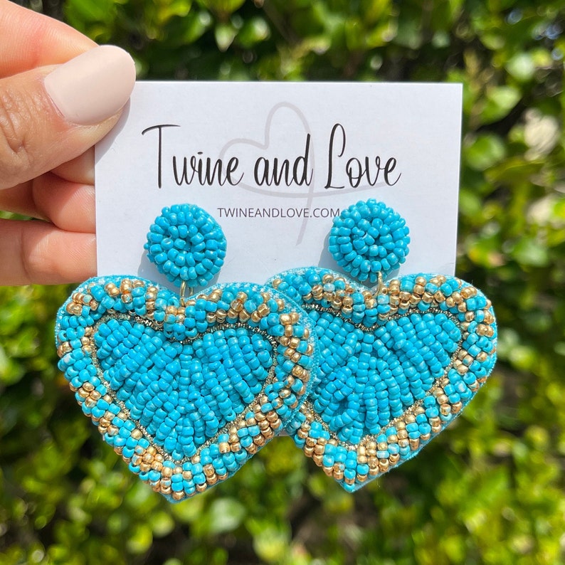 May include: Turquoise blue heart-shaped beaded earrings with gold accents. The earrings are large and dangle. The brand name "Twine and Love" and website "twineandlove.com" are printed on a white card behind the earrings.