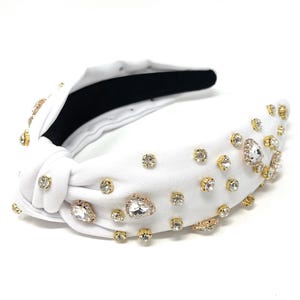 May include: White knotted headband with gold and clear rhinestones. The headband is decorated with various sizes of rhinestones, including some larger, teardrop-shaped stones.