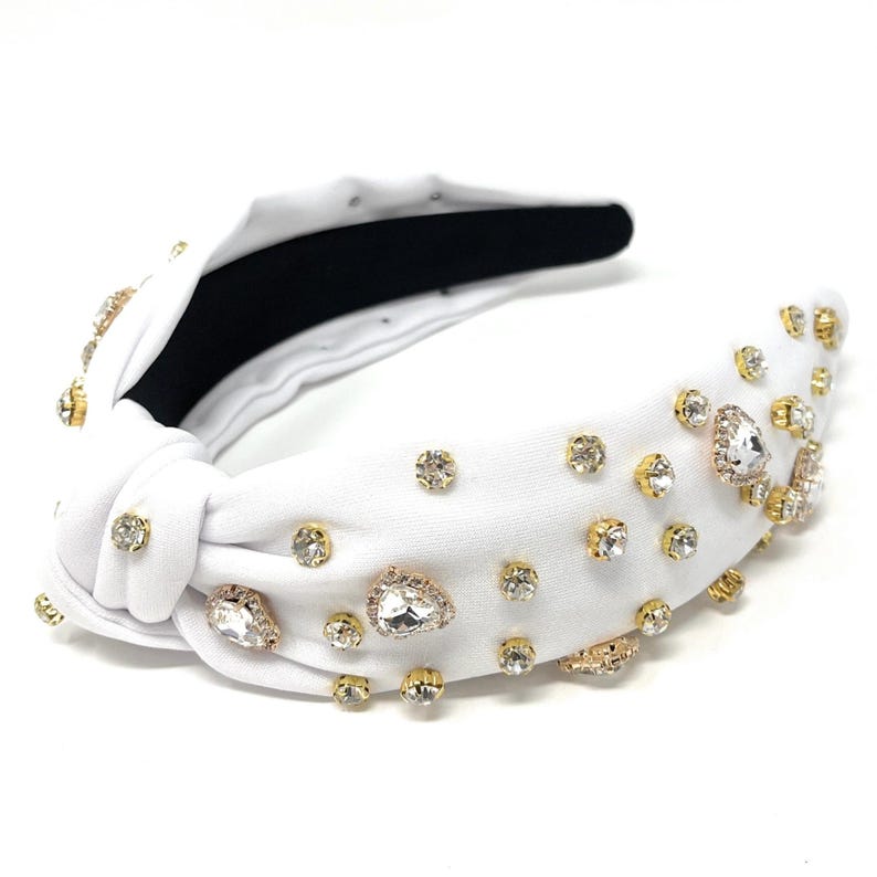 May include: White knotted headband with gold and clear rhinestones. The headband is decorated with various sizes of rhinestones, including some larger, teardrop-shaped stones.
