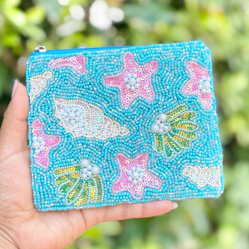 Hand holding a blue beaded pouch with starfish and shell designs against a blurred green background