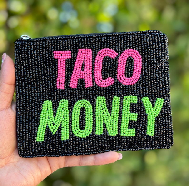 May include: Black beaded clutch with the words "TACO MONEY" in pink and green beads. The clutch has a zipper closure and is held in a hand.