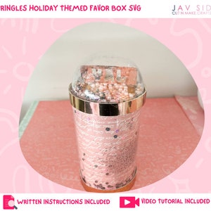 May include: A gold and clear plastic Pringles can favor box with a pink and white patterned design. The box is filled with pink and white glitter and beads. The text "PRINGLES HOLIDAY THEMED FAVOR BOX SVG" is at the top of the image. The text "JAV SID CUT N MAKE CRAFTS" is at the top right of the image. The text "WRITTEN INSTRUCTIONS INCLUDED" is at the bottom left of the image. The text "VIDEO TUTORIAL INCLUDED" is at the bottom right of the image.