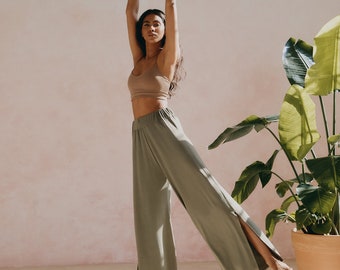 May include: A woman wearing a light brown tank top and olive green wide-leg pants with slits on the sides. She is standing with her arms raised above her head, looking at the camera. The background is a light pink wall with a potted plant.