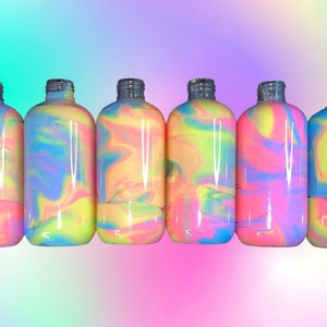 May include: A row of clear glass bottles filled with colorful, swirled liquid. The liquid features a mix of pink, yellow, blue, and purple hues. The bottles have silver-colored necks and are arranged against a gradient background.