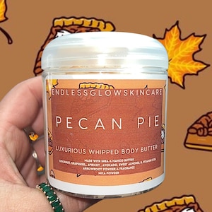 Puede incluir: Un tarro blanco con una etiqueta marrón que dice "ENDLESSGLOWSKINCARE PECAN PIE LUXURIOUS WHIPPED BODY BUTTER MADE WITH SHEA & MANGO BUTTER COCONUT, GRAPESEED, APRICOT, AVOCADO, SWEET ALMOND, & VITAMIN E OIL ARROWROOT POWDER & FRAGRANCE MICA POWDER". El tarro está en una mano que lo sostiene. El fondo es un patrón marrón con imágenes de pastel de nuez.
