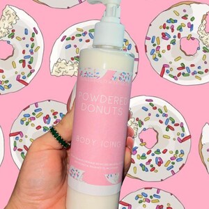 May include: A white bottle of body lotion with a pump dispenser, labeled "Powdered Donuts Body Icing." The bottle is held against a pink background patterned with illustrations of donuts with colorful sprinkles.