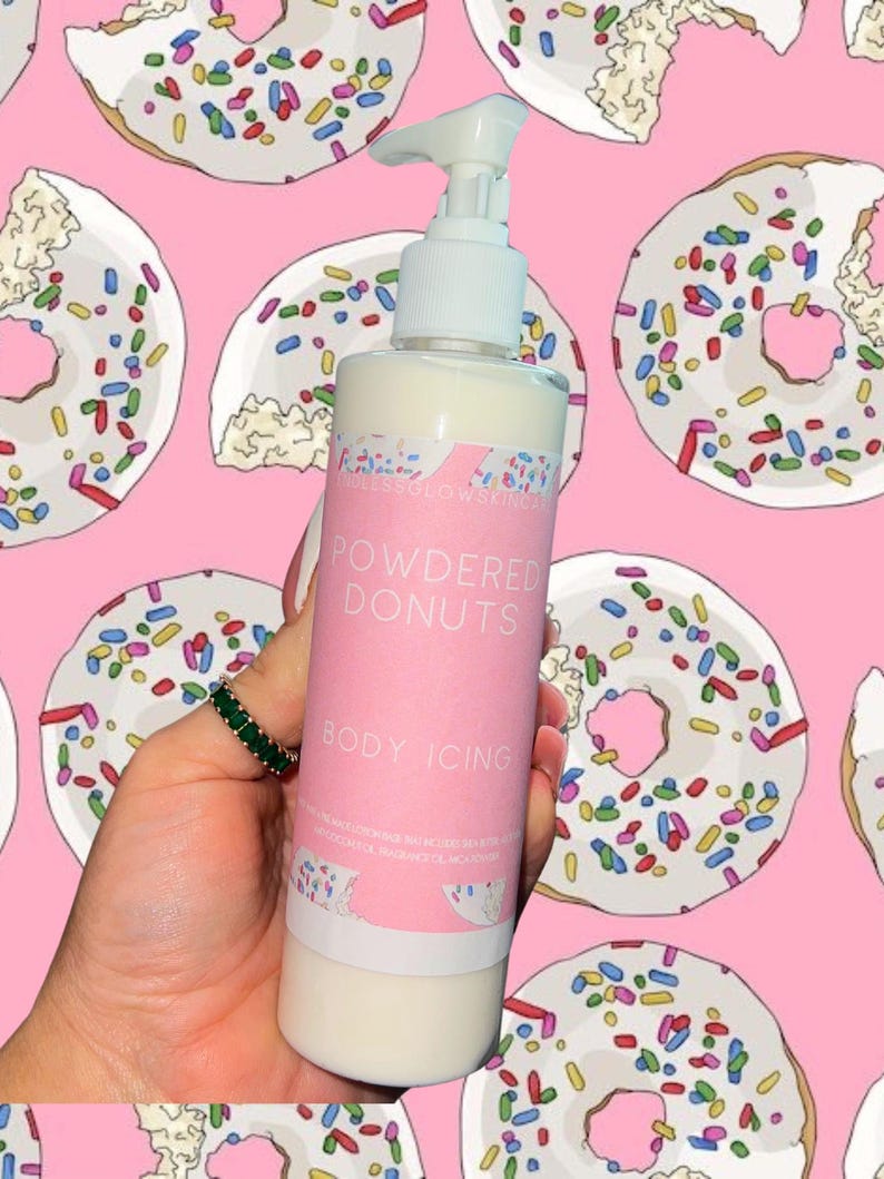 May include: A white bottle of body lotion with a pump dispenser, labeled "Powdered Donuts Body Icing." The bottle is held against a pink background patterned with illustrations of donuts with colorful sprinkles.
