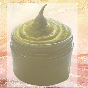 May include: A clear jar filled with a light yellow, whipped cream-like substance. The product has a swirl design and a peak at the top. The background is a mix of gold and pink tones.