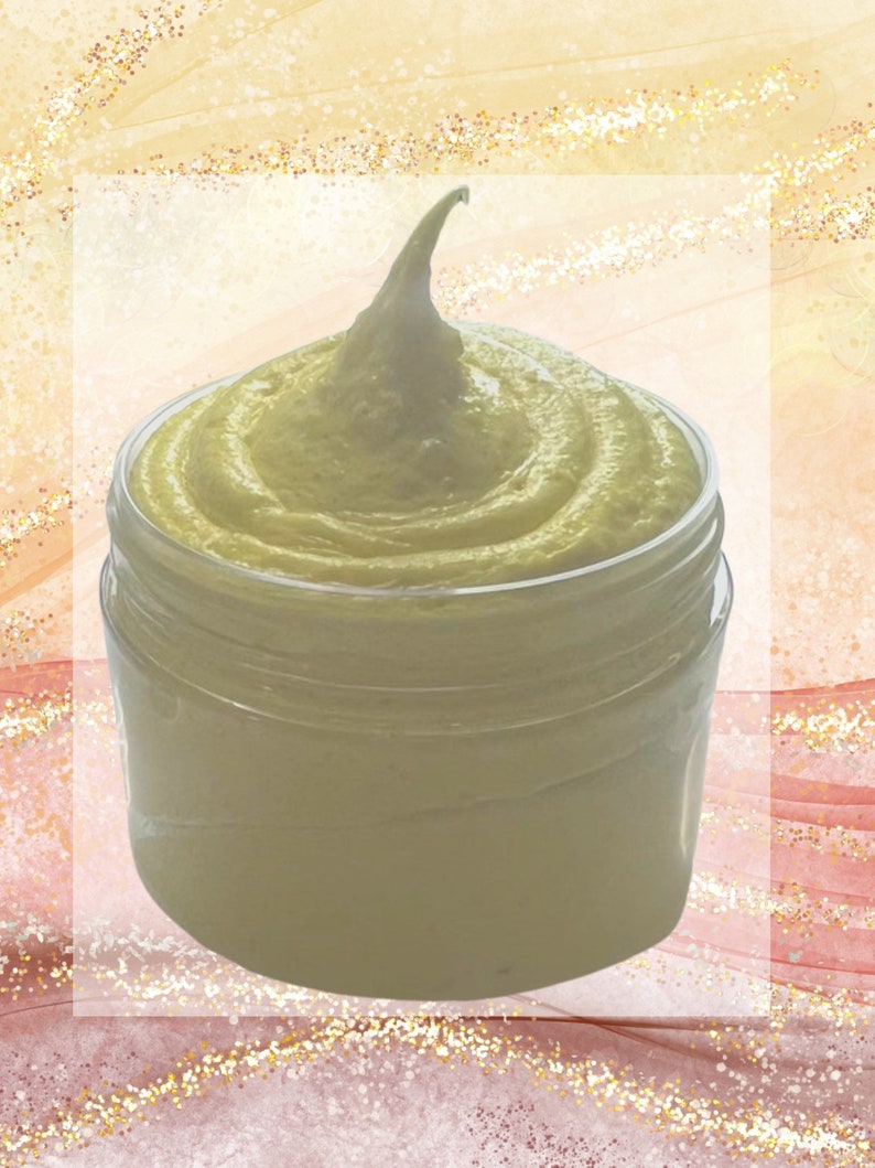 May include: A clear jar filled with a light yellow, whipped cream-like substance. The product has a swirl design and a peak at the top. The background is a mix of gold and pink tones.