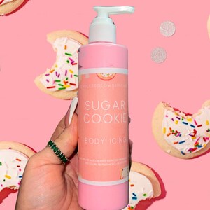 May include: A pink body lotion bottle with a white pump, labeled "Sugar Cookie Body Icing." The background features sugar cookies with white frosting and colorful sprinkles, along with silver confetti, all on a pink backdrop.
