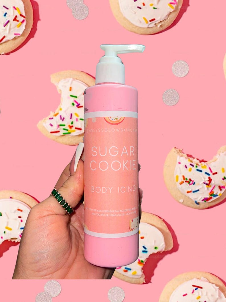 May include: A pink body lotion bottle with a white pump, labeled "Sugar Cookie Body Icing." The background features sugar cookies with white frosting and colorful sprinkles, along with silver confetti, all on a pink backdrop.