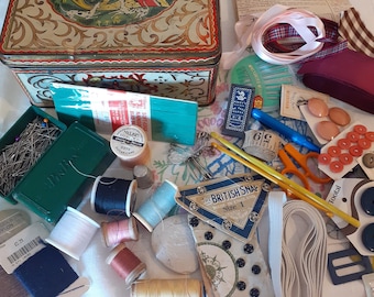 May include: A vintage tin box with a floral design and a scene of a horse and rider. The box is open and filled with various sewing supplies, including needles, thread, buttons, and elastic. The box is sitting on a white surface.
