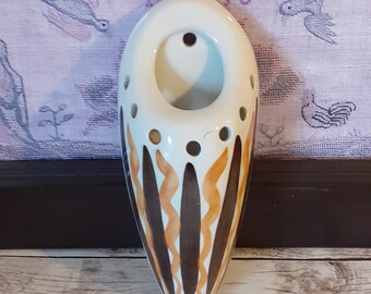 May include: A white ceramic wall pocket with a rounded top and a tapered bottom. It features brown and orange vertical stripes and circular cutouts. The wall pocket is designed for decorative purposes.