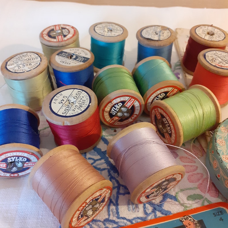 May include: A collection of vintage wooden spools of thread in various colors, including red, blue, green, and pink. The spools are labeled with the brand name "Sylko" and the number of yards of thread.
