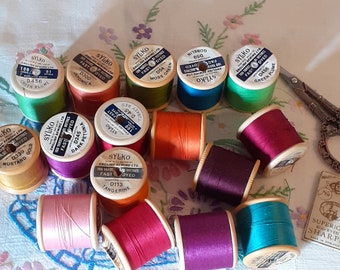 May include: A collection of colorful spools of Sylko thread in various shades, including green, orange, and purple, arranged on a floral patterned fabric. A pair of vintage scissors and a packet of needles are also present, suggesting a sewing or crafting theme.