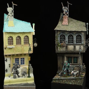 May include: Two miniature buildings with detailed facades, one painted in a light brown with a blue roof and the other in a dark brown with a green roof. Both buildings have a clock on the side and a figure on the roof. The buildings are on a base with a cobblestone street and a figure standing in front of each building.