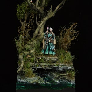 May include: A miniature figure of a wizard standing in a forest setting. The wizard is wearing a blue and green robe and holding a staff. The forest is depicted with a small stream and a tree with green leaves.