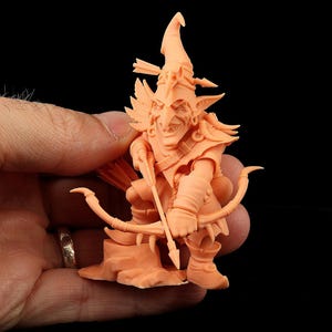 Op de afbeelding: Een perzikkleurig miniatuurfiguurtje van een goblin-boogschutter met een puntmuts voorzien van een pijl en schedeldetail. Het personage heeft puntige oren, een brede grijns en houdt een boog en pijl vast op een kleine rotsachtige basis.