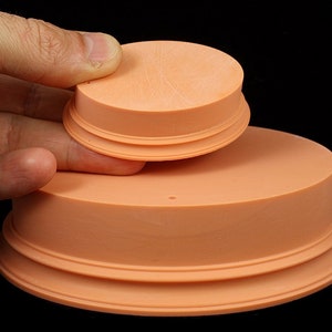 Könnte beinhalten: Zwei pfirsichfarbene, kreisförmige Objekte mit einem abgestuften Design. Das kleinere wird über dem größeren gehalten. Die Objekte haben eine glatte Oberfläche mit einigen sichtbaren Kratzern. Der Hintergrund ist einfarbig schwarz.