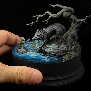 Op de afbeelding: Een miniatuur diorama met een wolf en een persoon bij een vijver. De wolf is donkergrijs en de persoon draagt groen. De vijver heeft waterlelies en een boom op de achtergrond. De basis is zwart.